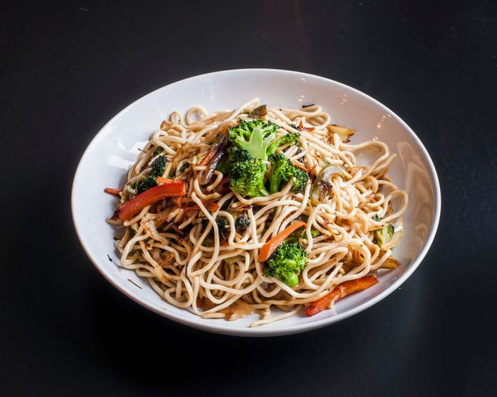 Een kom noedels met broccoli en paprika op een zwarte ondergrond.