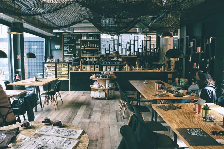 Een koffiezaak met lange houten tafels, gasten aan het raam en de toonbank achterin.