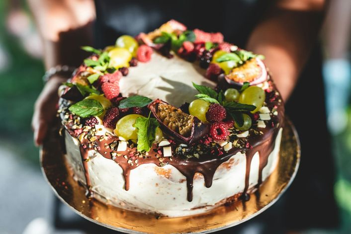 Two hands hold a cake topped with berries, grapes, mint and a chocolate drip.