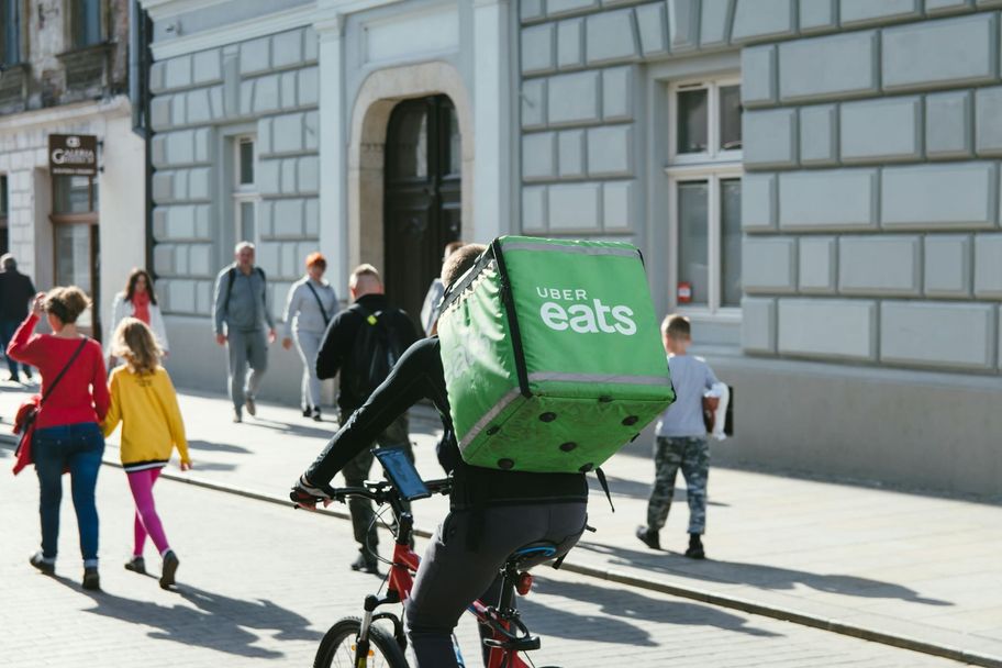 Een fietskoerier met een grote groene bezorgbox rijdt door een winkelstraat vol voetgangers.