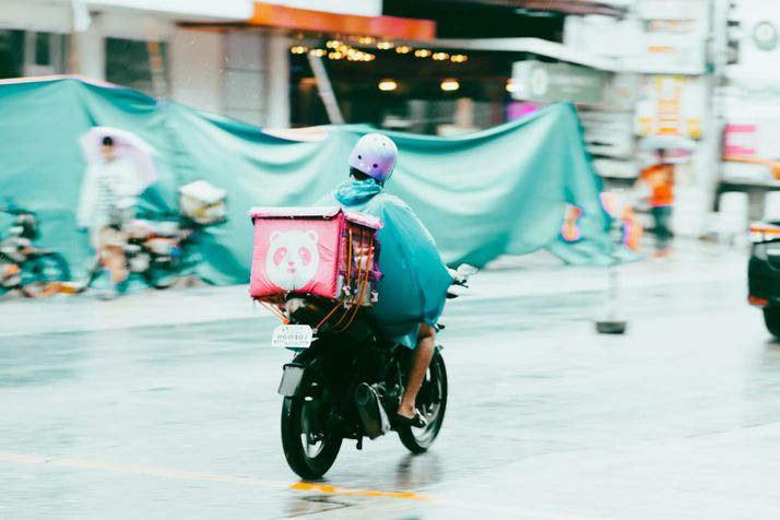 Een bezorger met een roze bezorgbox rijdt op een scooter door een natte straat.