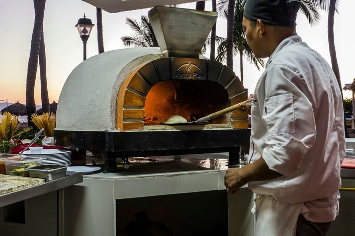 Een kok schuift een pizza in een witte houtoven, buiten in de schemering.