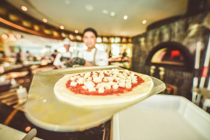 A pizza chef holds a peel with a topped pizza, in front of a wood oven.