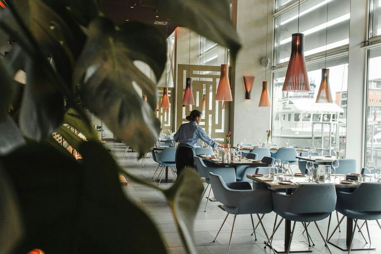 Een medewerker dekt tafels in een lichte eetzaal met blauwe stoelen en hanglampen.