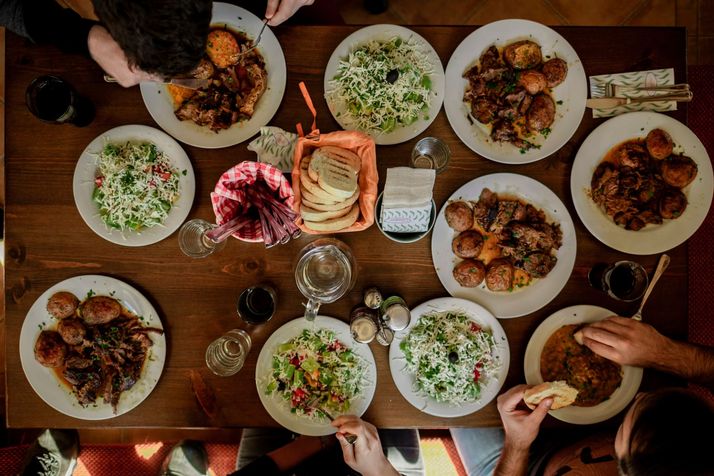 Een tafel van boven gezien, vol schalen vlees, salade en brood, met gasten die opscheppen.