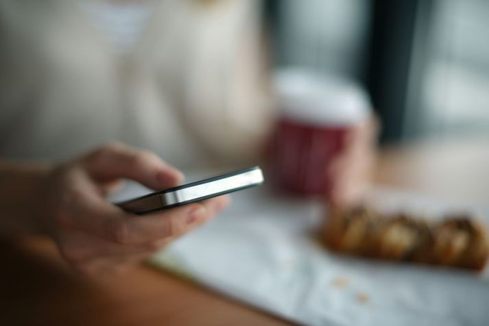 Een hand met een telefoon aan tafel, naast een beker afhaalkoffie en een broodje.