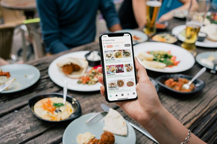 Een hand houdt boven een volle tafel een telefoon vast met een app vol gerechten.