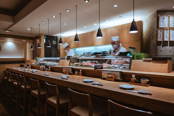 Twee sushikoks achter een vitrine, met lege krukken langs de toonbank.