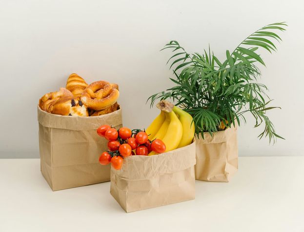Drie papieren tassen op een lichte tafel, gevuld met gebak, tomaten, bananen en groen.
